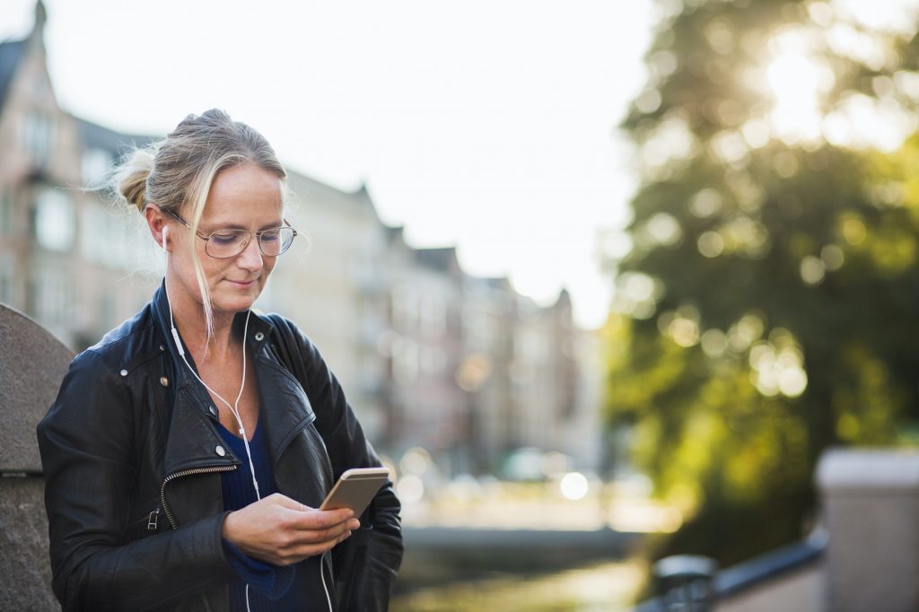 Mature woman with earphones looking at smart phone while standing at street