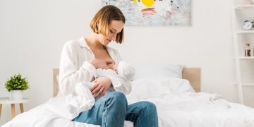 Mother in jeans sitting on bed and breastfeeding baby