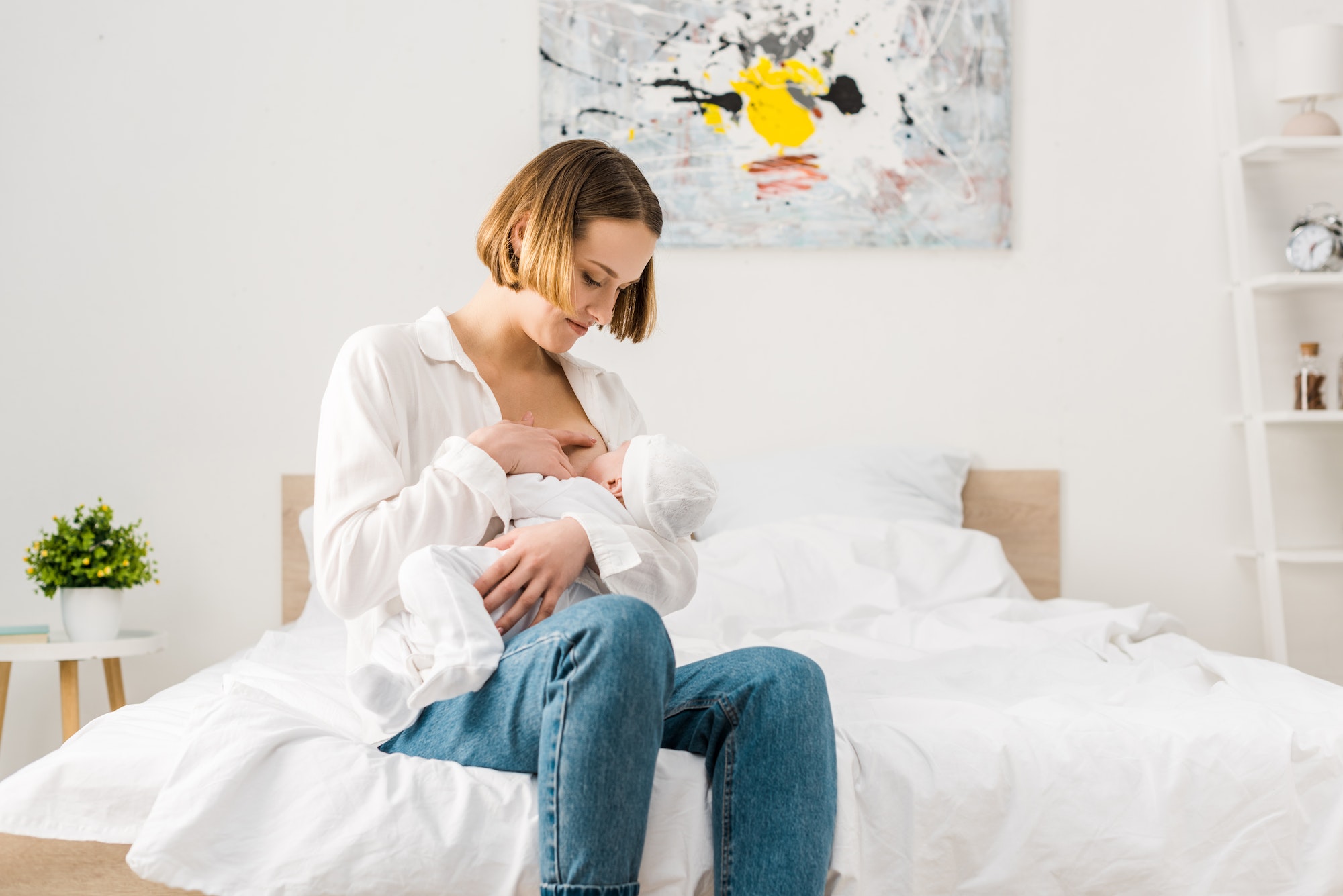 Mother in jeans sitting on bed and breastfeeding baby