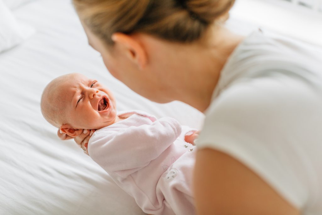 Young woman cradling crying baby daughter on bed, over shoulder view