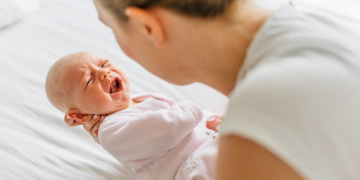 Young woman cradling crying baby daughter on bed, over shoulder view