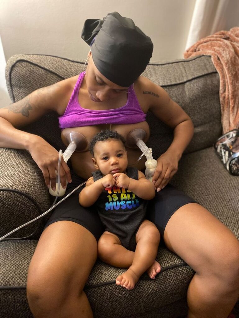 Image of black mum sitting on a chair and using double electric pump with baby nestled in between her legs